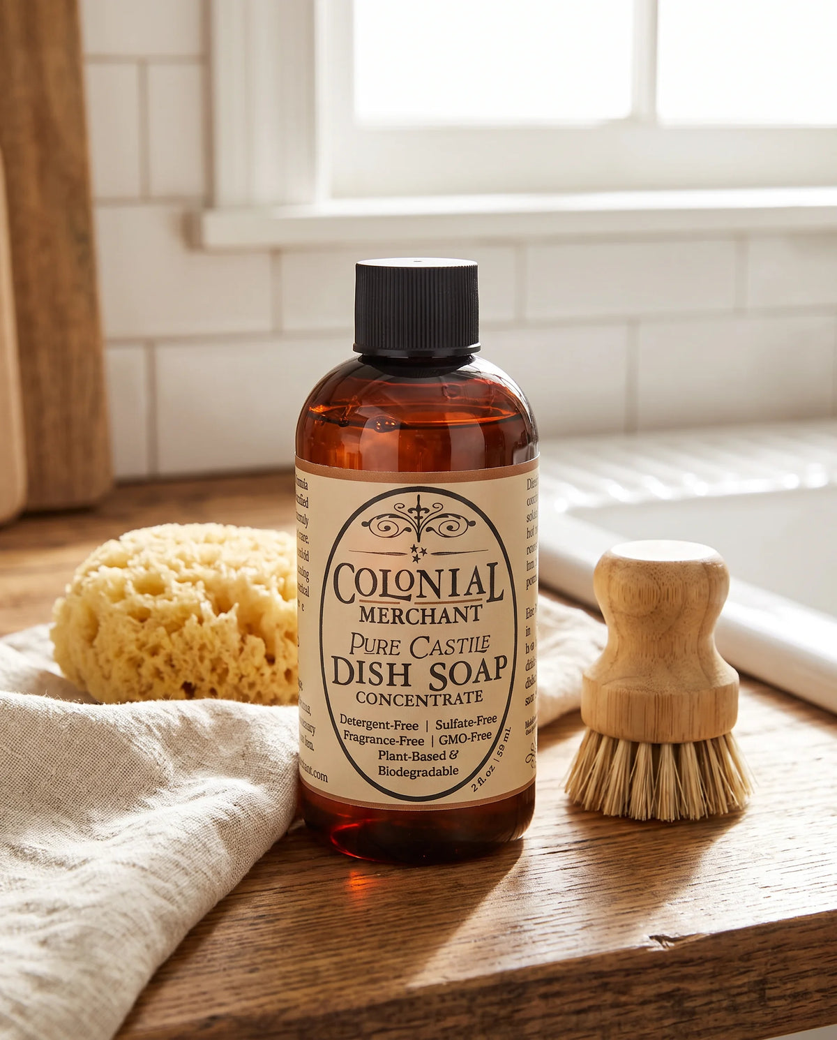 A bottle of Colonial Merchant Pure Castile Dish Soap Concentrate stands on a wooden surface in a bright bathroom setting, with a sponge, cloth, and wooden brush nearby.