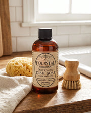 A bottle of Colonial Merchant Pure Castile Dish Soap Concentrate stands on a wooden surface in a bright bathroom setting, with a sponge, cloth, and wooden brush nearby.