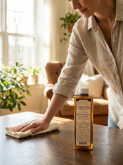Person cleaning a wooden surface with a bottle of Cornish Pure Wood Polish in a home setting.